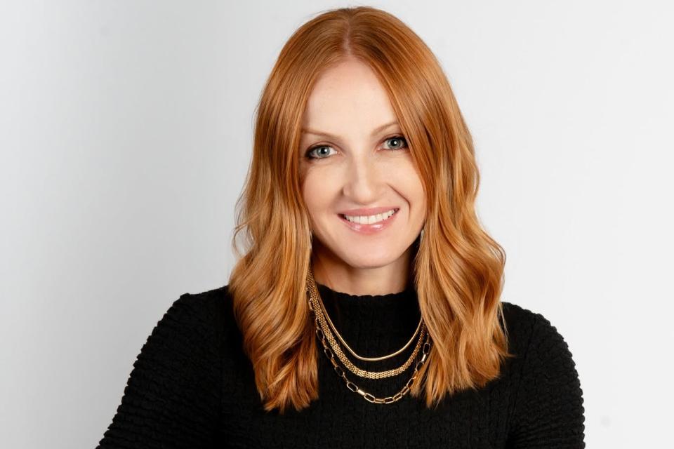 Woman with long red hair wearing a black textured top and layered gold necklaces, seated and smiling against a plain light background.