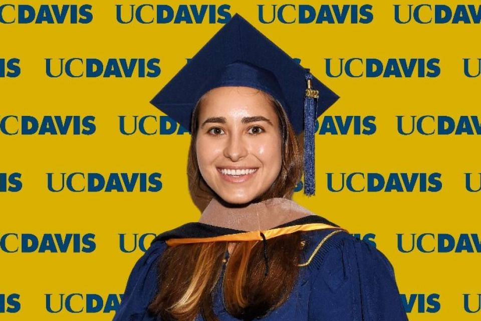 Maria Ahumada wearing a blue graduation cap and gown stands in front of a yellow UC Davis backdrop with repeated logos.