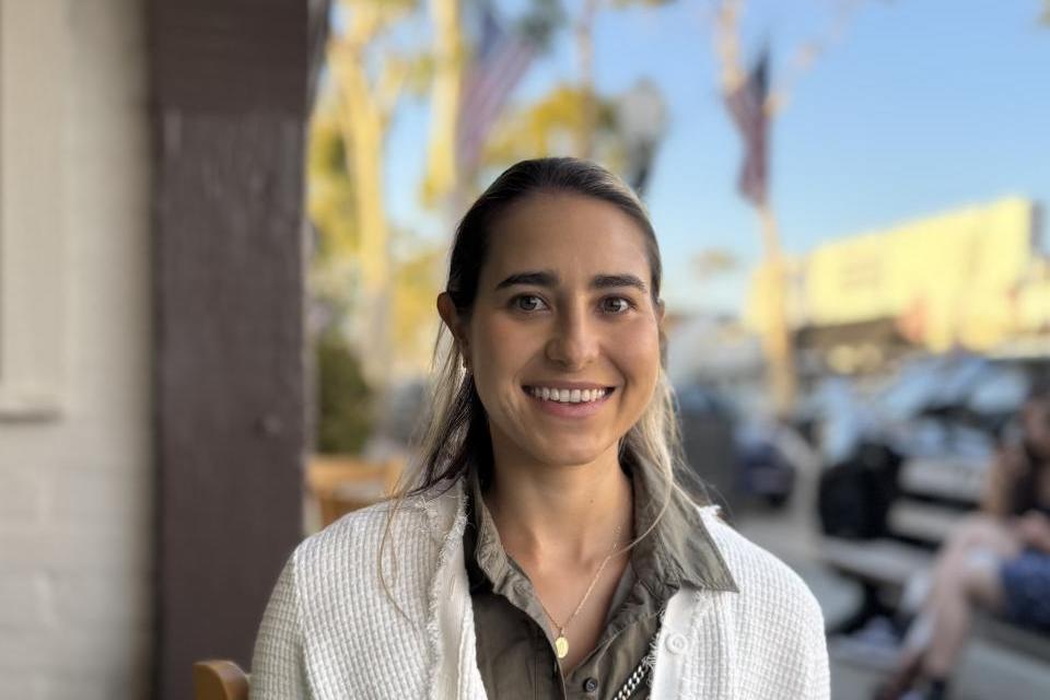 A woman with long hair, wearing a white sweater over a green shirt, sits outdoors at a restaurant table, smiling at the camera. Trees and parked cars are visible in the background.