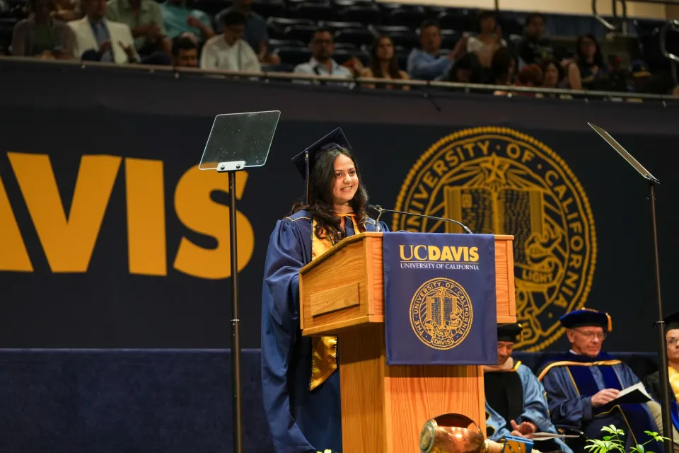 A student in cap and gown speaks at a podium during a UC Davis graduation ceremony, with faculty seated behind and audience in the background.