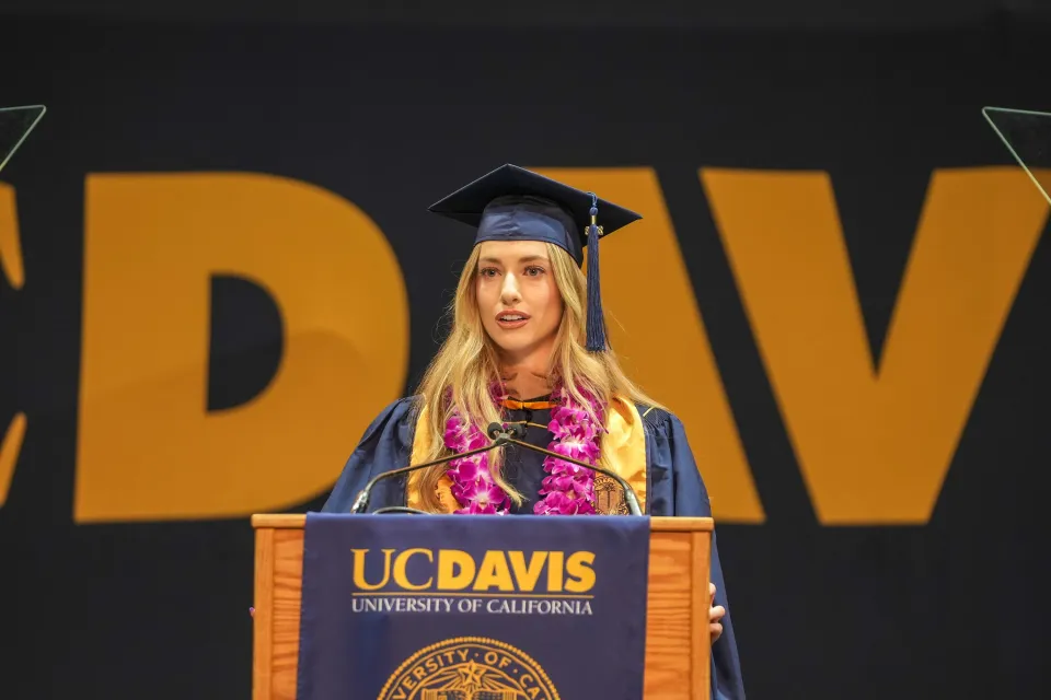 A graduate in cap and gown speaks at a podium with a UC Davis sign, wearing a purple lei, during a commencement ceremony.