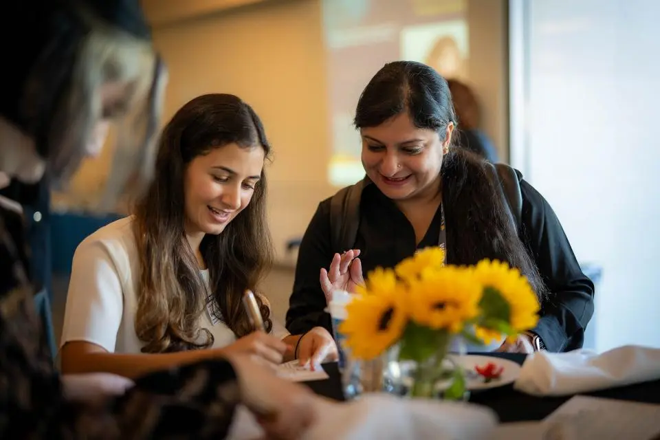 Two women sit at a table with sunflowers, smiling and writing on paper during an indoor event.