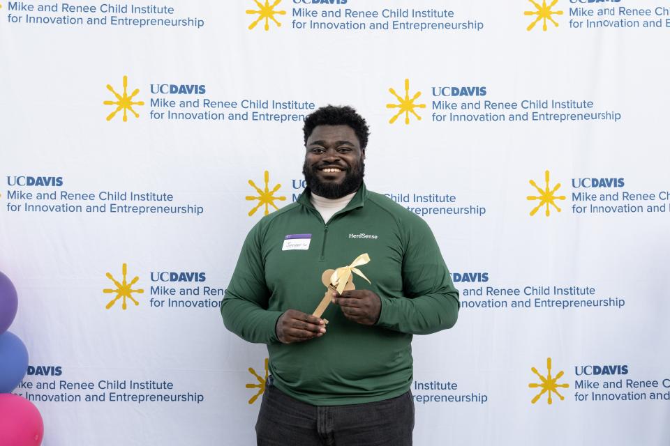 Jeremy Twumasi Ampofo in a green polo shirt holds a wooden award in front of a UC Davis backdrop.