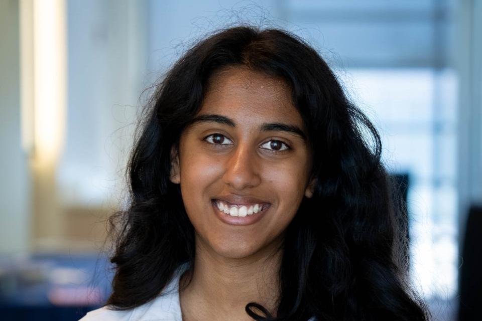 Rachel Johnson with long curly hair, wearing a white ruched blouse, stands indoors and smiles at the camera. The background is out of focus.