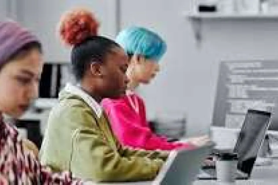 Three people are sitting in a row at desks, working on laptops in a modern classroom or office setting. Computer code is visible on a monitor in the background.
