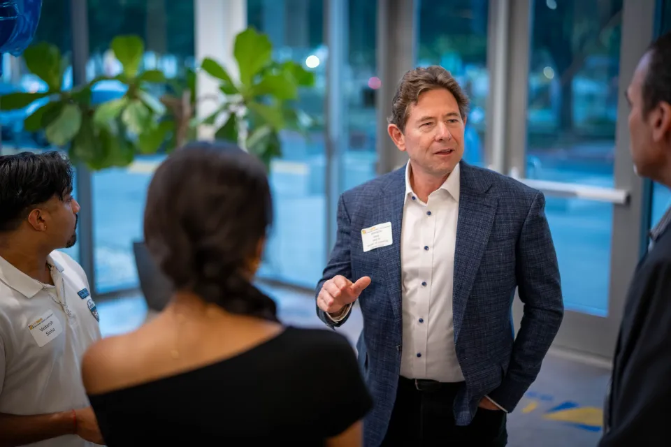 A man in a suit jacket talks to three people at an indoor event, wearing a name tag, with large windows and plants in the background.