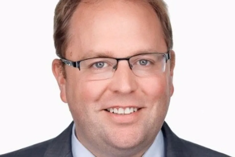 Man with short light brown hair, glasses, and a gray suit jacket over a light blue shirt and patterned tie, smiling at the camera against a plain white background.