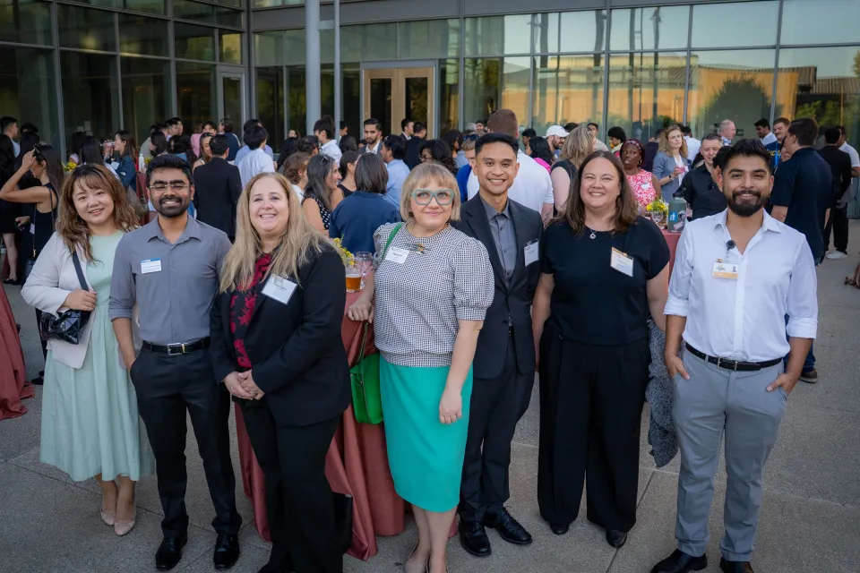 A group of seven people pose for a photo outdoors at a formal event, with other attendees and tables visible in the background.