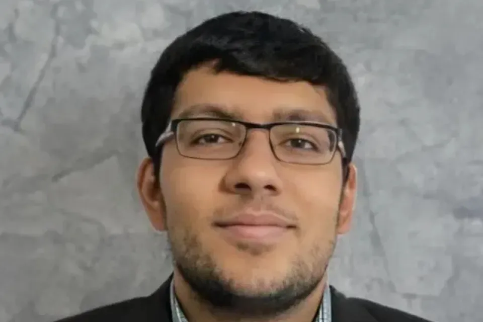 A man wearing glasses and a checked shirt with a dark blazer is posed in front of a gray textured background.