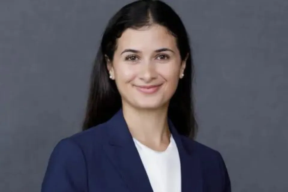 Woman with long dark hair wearing a navy blazer and white blouse, smiling in front of a plain gray background.