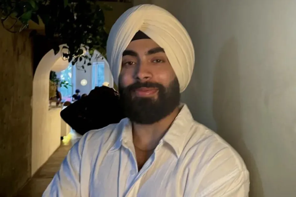 A person wearing a white turban and white shirt stands indoors with arms crossed, facing the camera and smiling slightly.