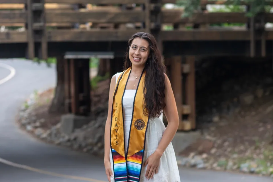 Woman in a white dress and graduation stole stands on a road in front of a wooden bridge, smiling at the camera.