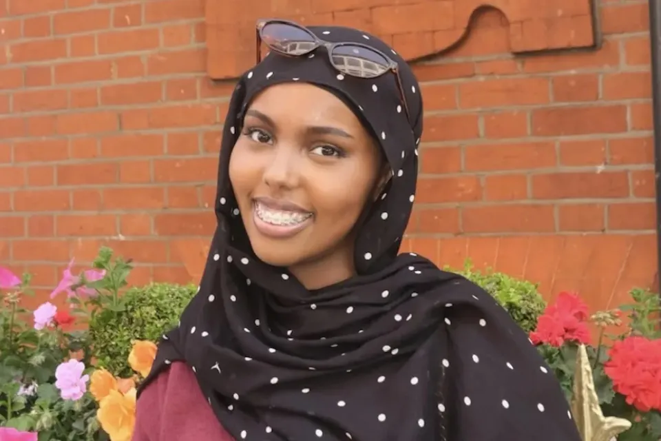 A woman wearing a black and white polka dot hijab and sunglasses on her head smiles in front of a brick wall and colorful flowers.