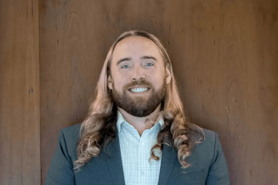 A person with long blond hair and a beard, wearing a suit jacket and checked shirt, stands smiling in front of a wooden background.