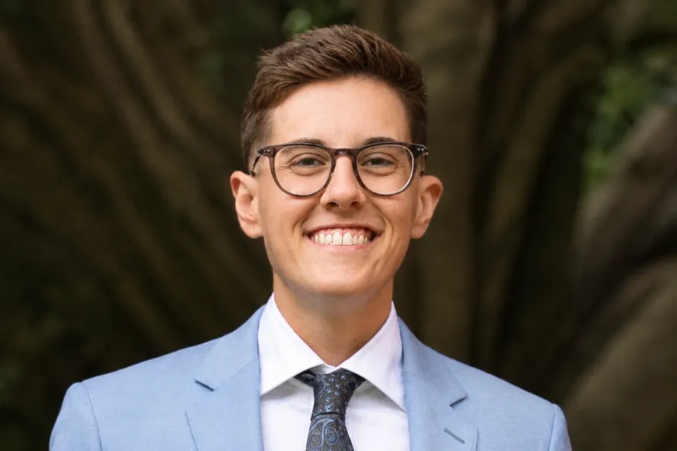 A person wearing glasses and a light blue suit jacket smiles at the camera, standing outdoors with a tree trunk in the blurred background.