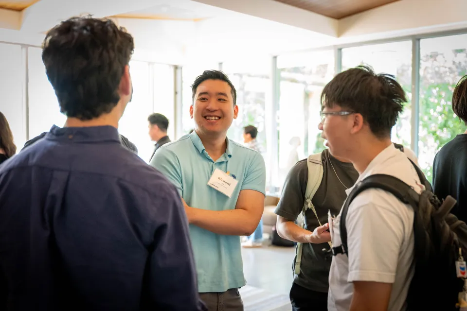 Three people are standing and talking at an indoor event. One person in a light blue shirt is smiling, while the others listen. There are more people in the background.