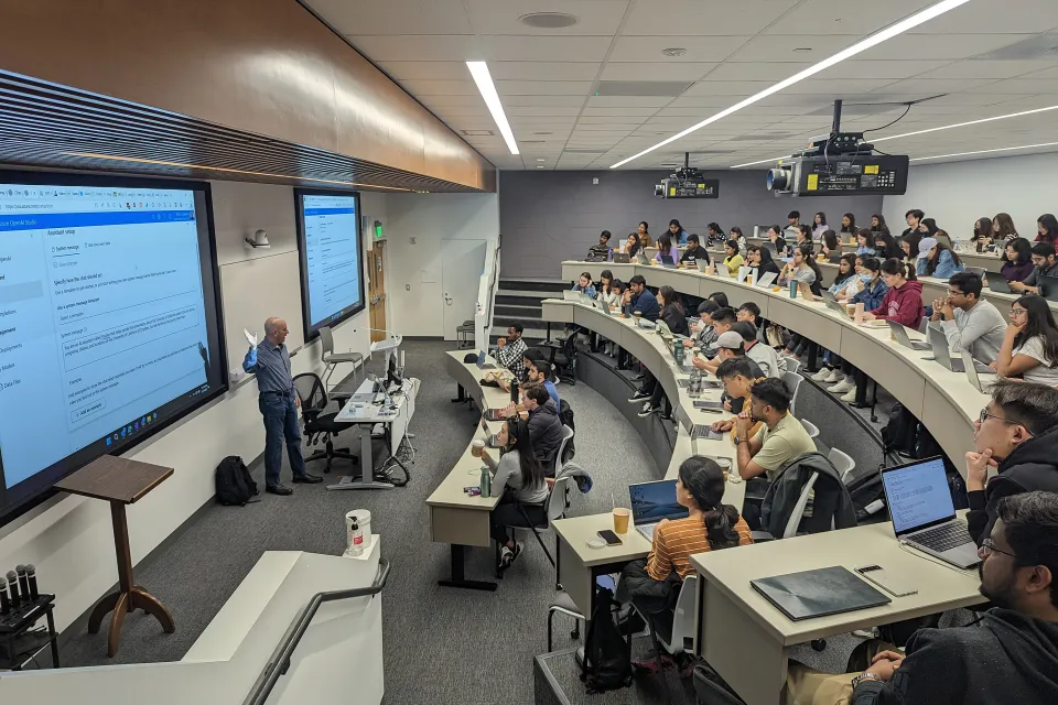 A professor stands at the front of a tiered lecture hall, presenting slides to students seated at desks with laptops and notebooks.