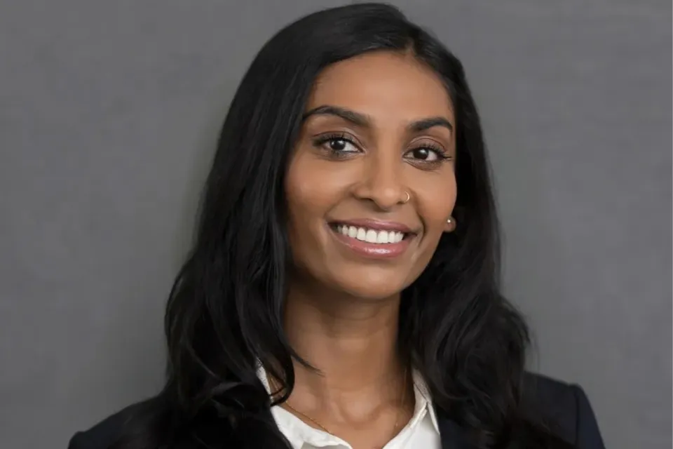 Woman with long dark hair, wearing a white shirt and dark blazer, smiling in front of a plain gray background.