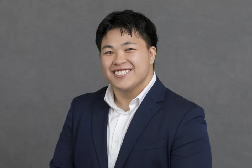 Person in a navy suit and white shirt smiling in front of a plain gray background.