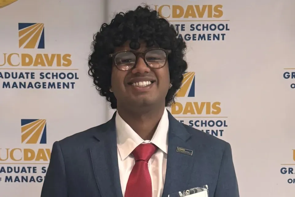 Person in a suit, red tie, and glasses smiling in front of a UC Davis Graduate School of Management backdrop.