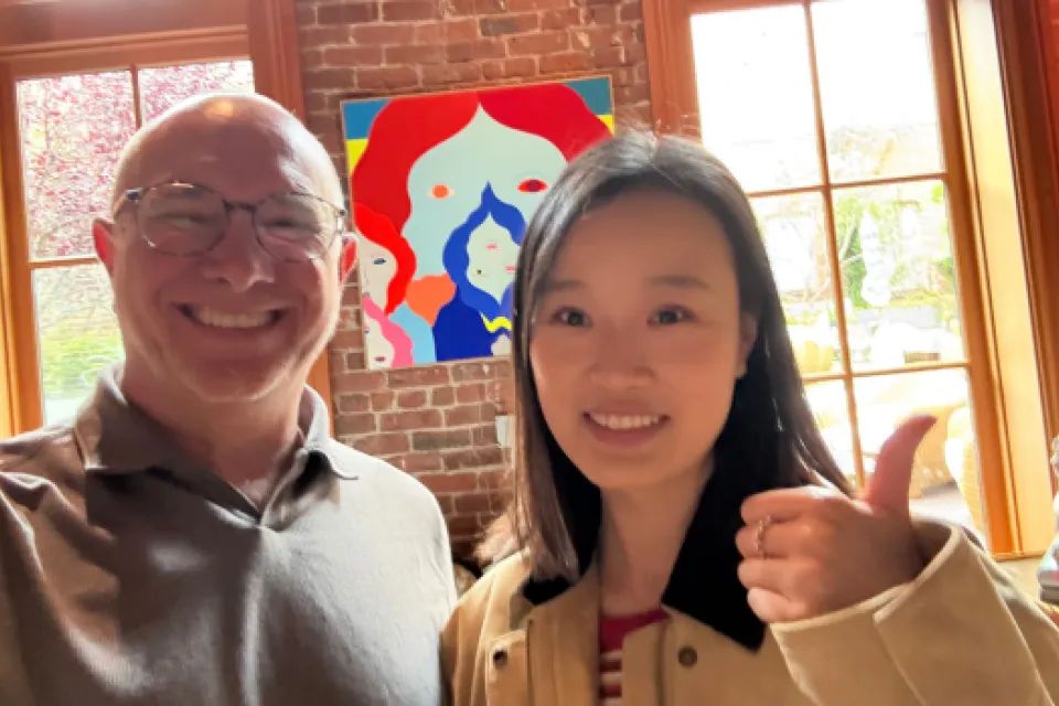 Two people smile at the camera indoors; one gives a thumbs up. A colorful abstract painting and large windows with a garden view are in the background.