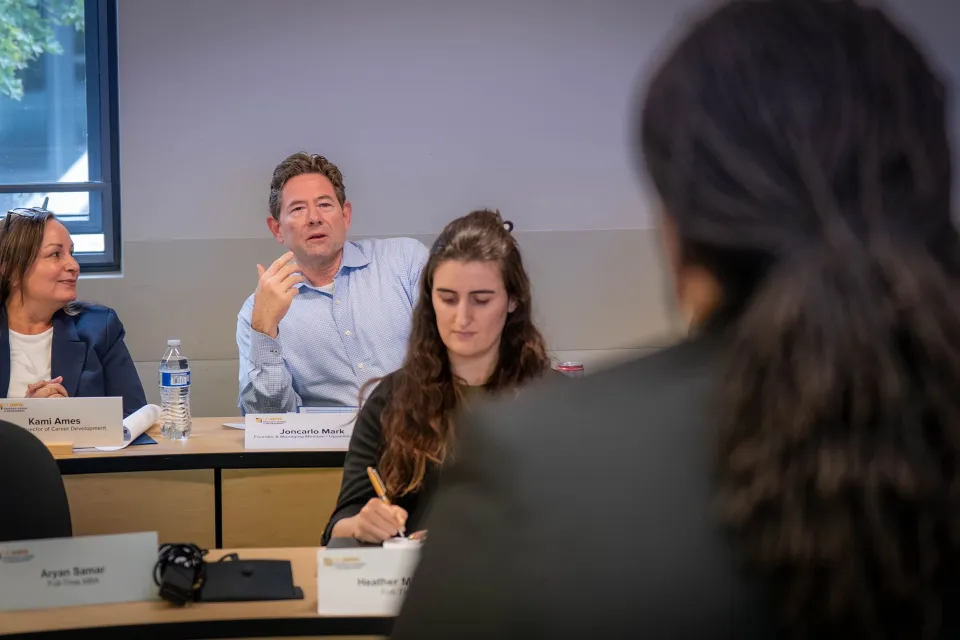 A man speaks during a classroom discussion while two women listen and take notes. Nameplates and notebooks are on the desks.