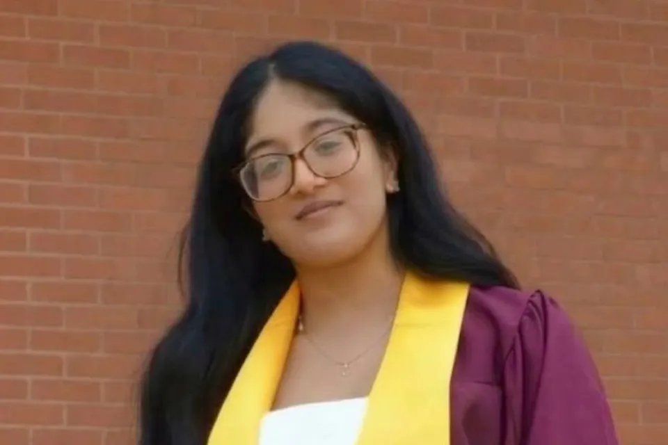 A young woman with long dark hair and glasses wearing a maroon graduation gown and yellow stole in front of a brick wall.