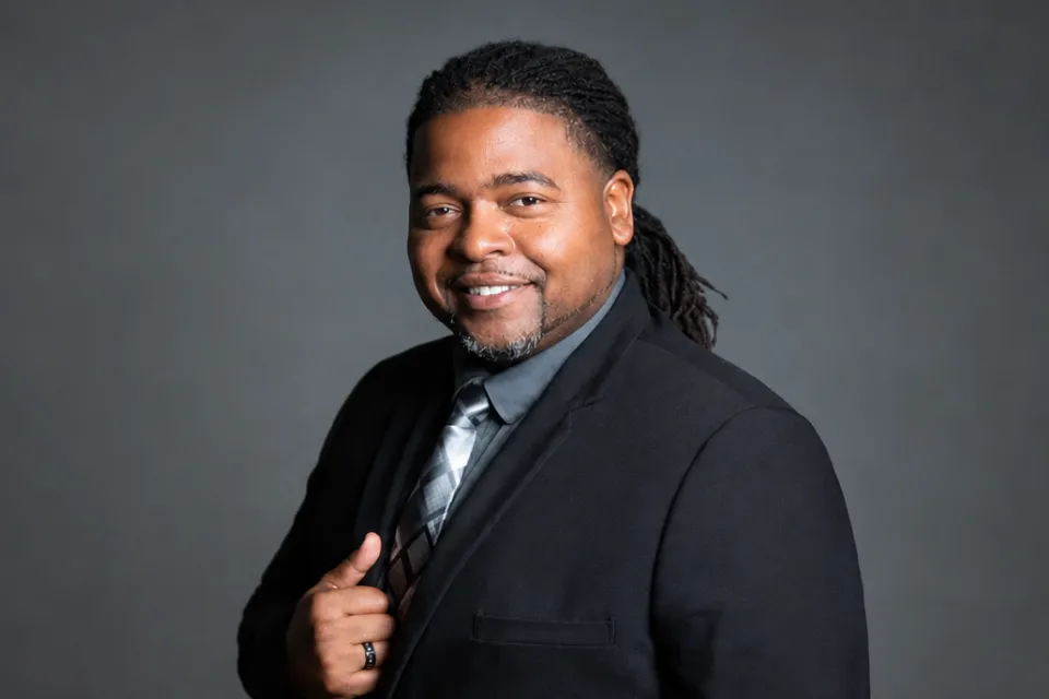 A man in a black suit, grey shirt, and patterned tie smiling at the camera.