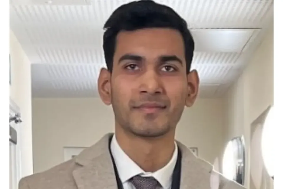 A professional headshot of a man in a beige blazer, white shirt, and tie.
