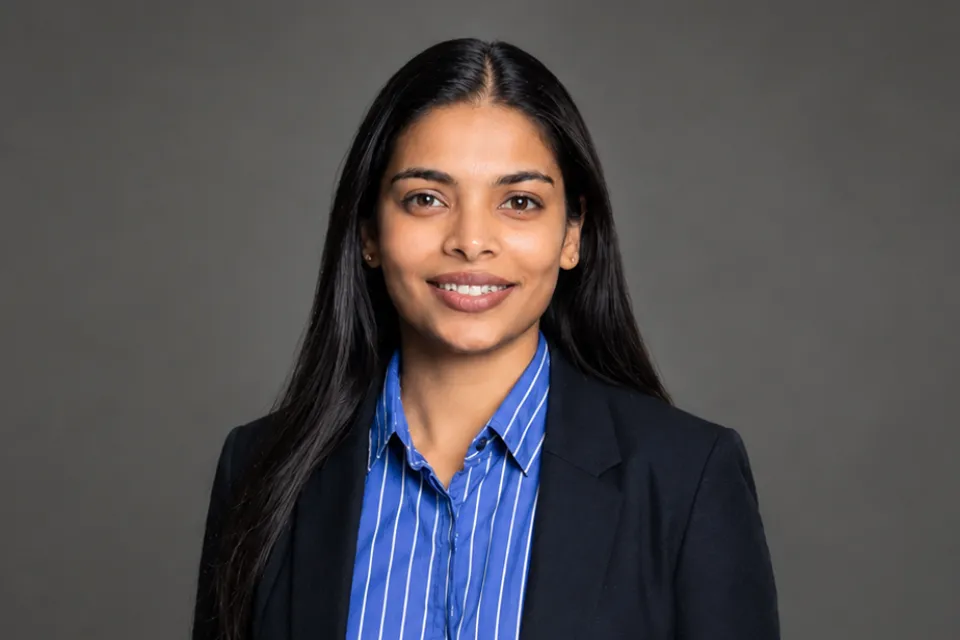 Professional portrait of a woman with long dark hair wearing a blue striped shirt and a black blazer against a grey background.
