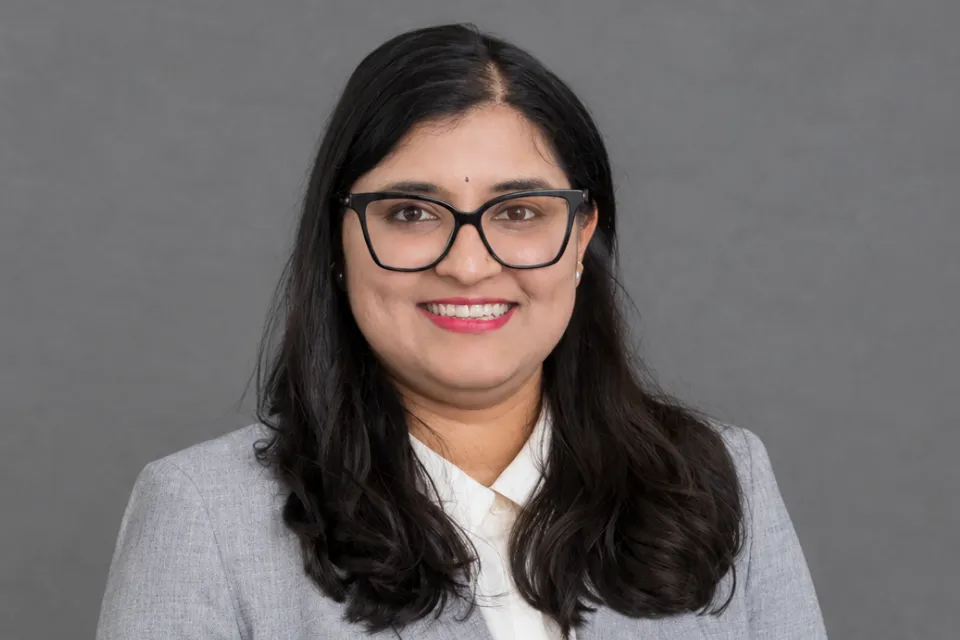 A professional headshot of a smiling woman with dark hair and glasses wearing a grey blazer over a white collared shirt.