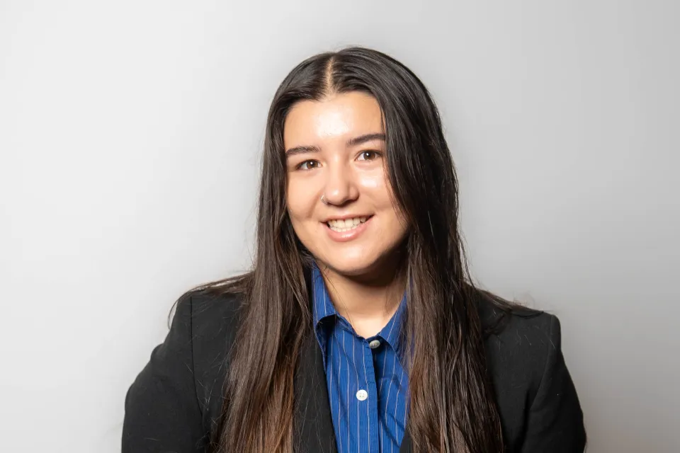 A young woman with long dark hair wearing a black blazer over a blue pinstriped button-down shirt.