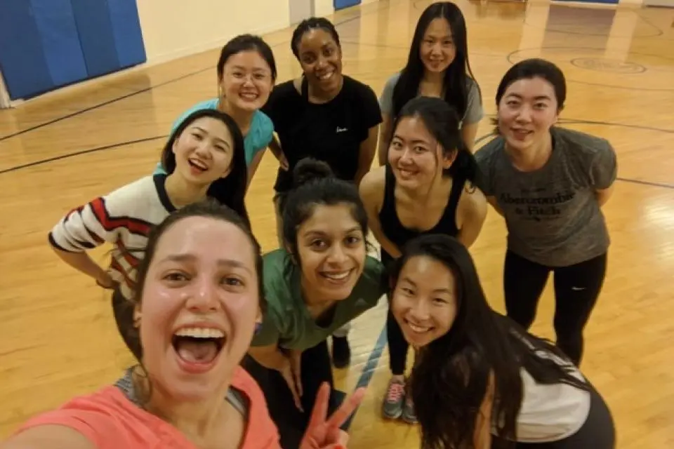 Nine women in athletic wear pose and smile for a group selfie in a gym with a wooden floor and blue wall padding.
