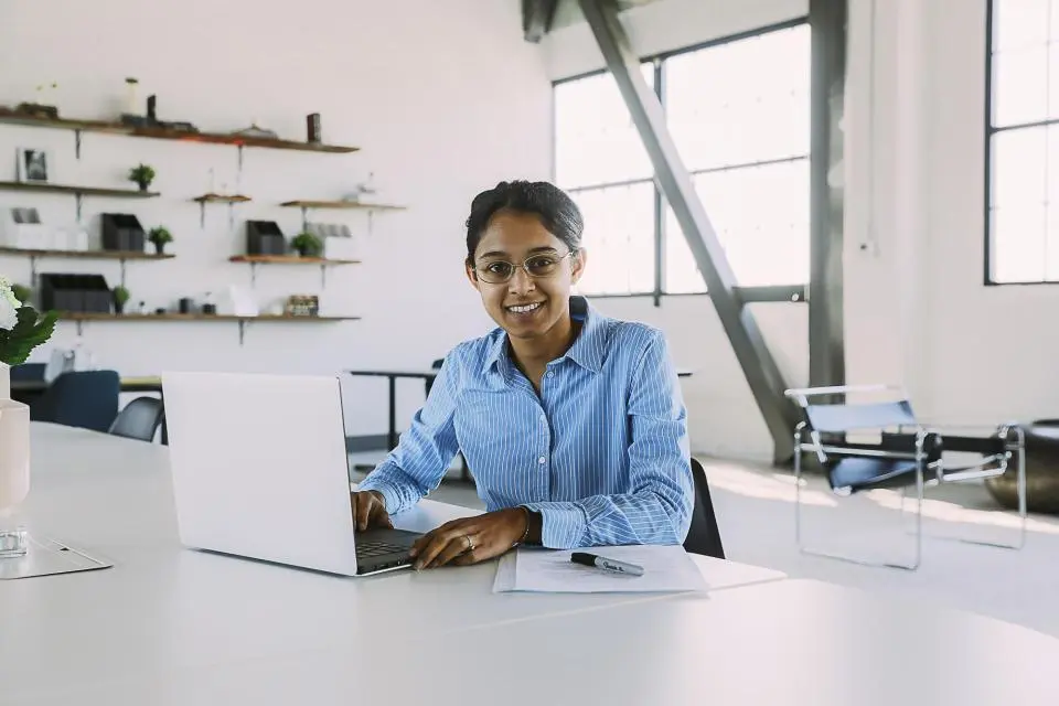 A woman wearing glasses sits at a desk with a laptop and documents in a bright, modern office space.