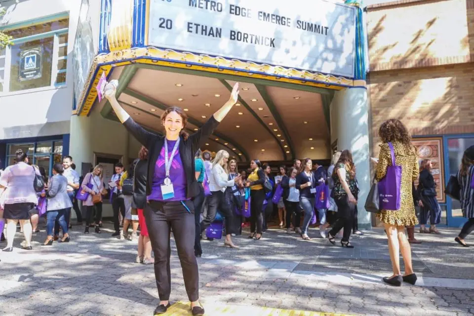 A woman stands smiling with arms raised in front of a theater entrance, surrounded by a crowd of people, under a marquee sign displaying event information.