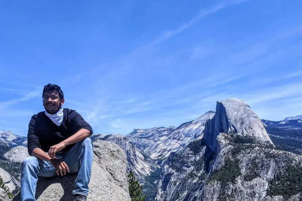 A man sits on a large rock overlooking a mountainous landscape with clear blue sky and the distinct peak of Half Dome in the background.