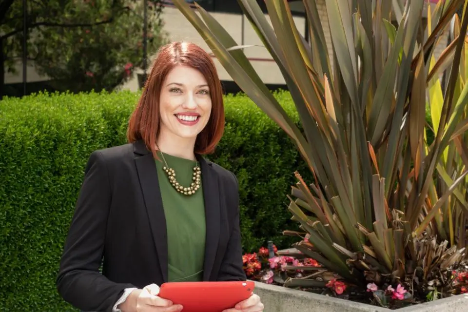 A woman with red hair, wearing a black blazer and green blouse, sits at an outdoor table holding a red tablet, with greenery and plants in the background.