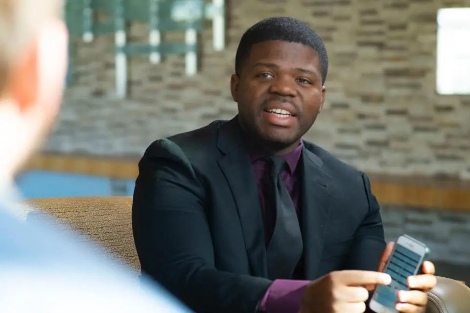 A man in a suit holding a smartphone sits on a couch, talking to another person whose back is to the camera.