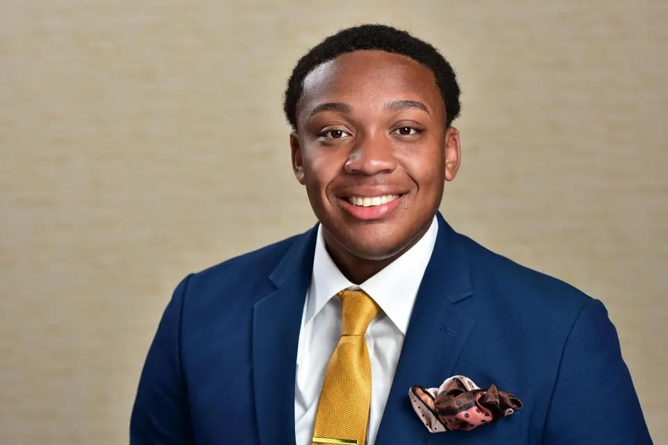 A man in a blue suit, white shirt, and yellow tie is smiling at the camera with a beige background behind him.