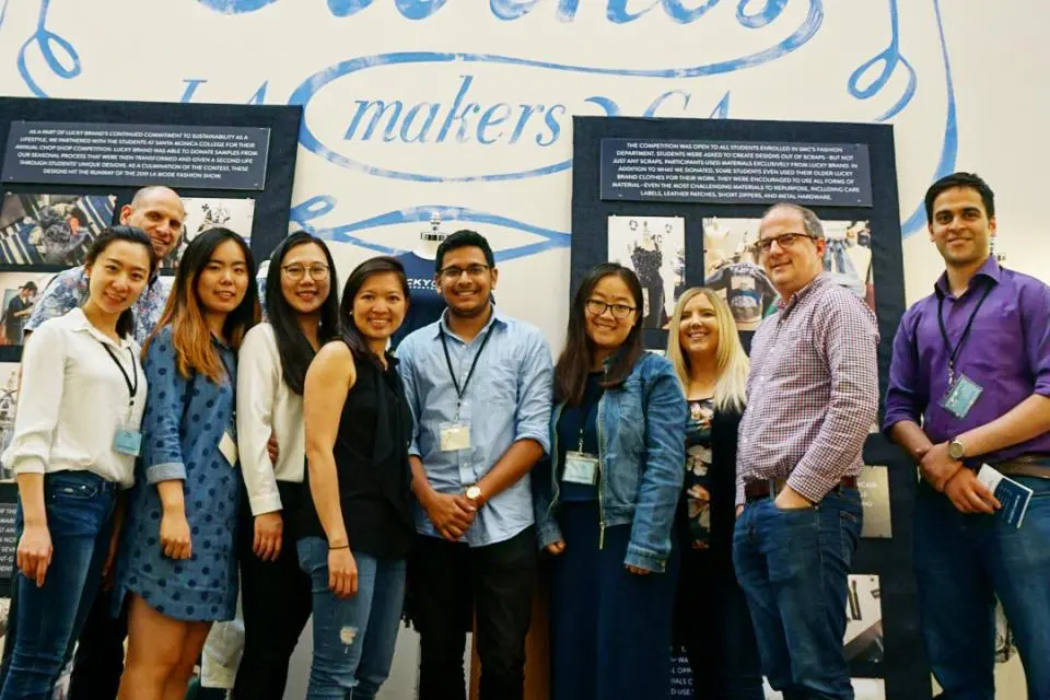 A group of eleven people pose and smile in front of a large wall sign reading "Lucky Brand House of Fine Clothes.