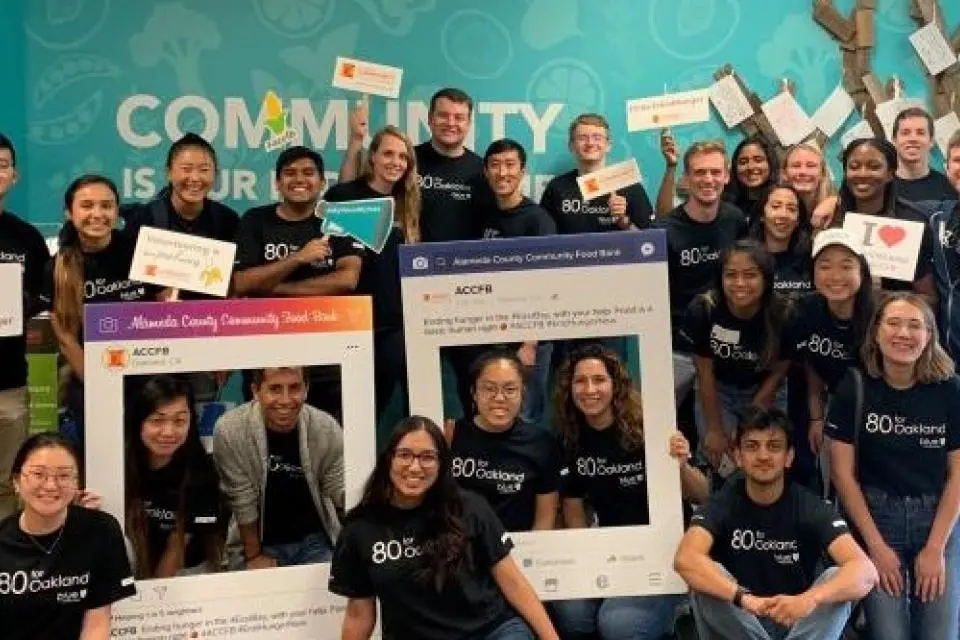 A diverse group of people in matching "80 Oakland" shirts pose together indoors, holding signs and photo frames, in front of a teal wall with "Community" written on it.